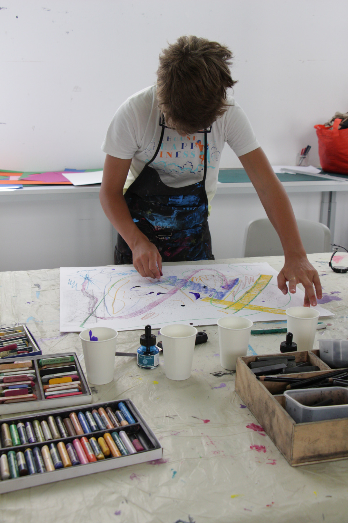 Face à l'objectif, un pré-adolescent dessine sur une feuille. Devant lui, des boîtes de crayons, de feutres et de peinture.
