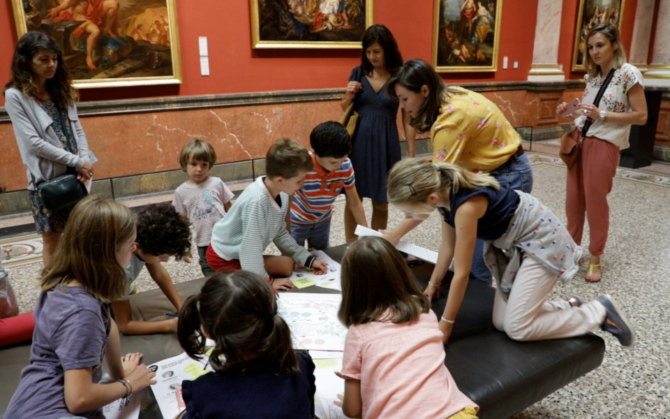 Une médiatrice reçoit des familles dans la galerie des colonnes du musée Fabre. Les enfants s'appuient sur des bancs pour utiliser le livret de visite.