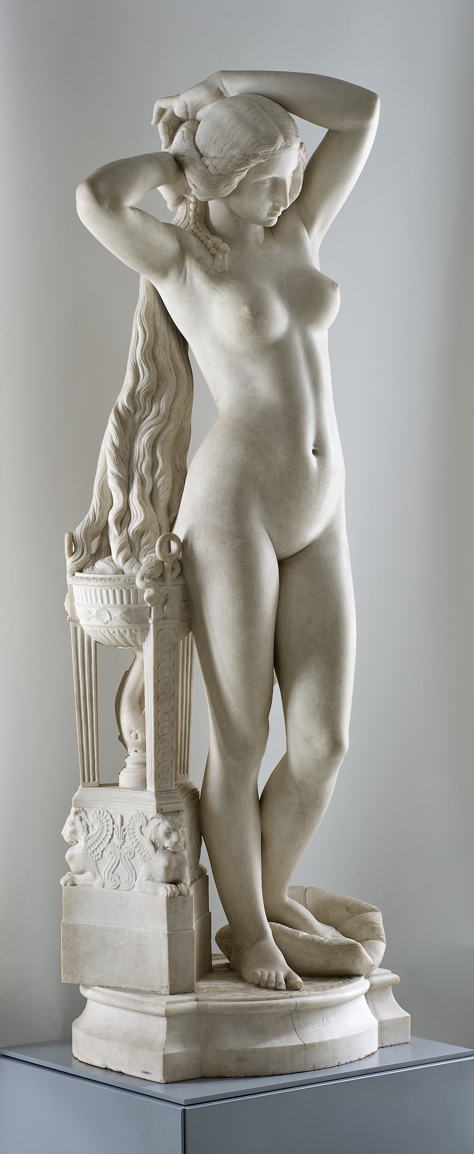 Sculpture en marbre représentant une jeune femme nue et debout (Nyssia, l'épouse du roi Candaule) et se tenant l'arrière des cheveux de ses deux mains, et le pied gauche appuyé sur un coussin.