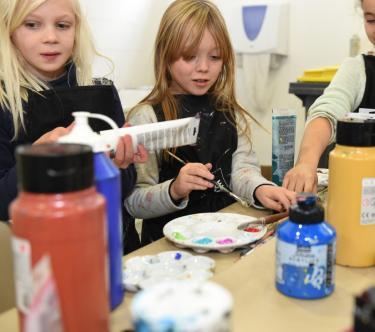 Trois fillettes sont en train de peindre sur une table. Au premier plan, les pots et tubes de peinture.
